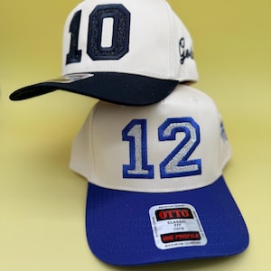 Puede incluir: Dos gorras de b&eacute;isbol apiladas verticalmente. La gorra superior es de color blanco roto con visera negra y el n&uacute;mero 10 en purpurina negra. La gorra inferior es de color blanco roto con visera azul y el n&uacute;mero 12 en purpurina azul, con una pegatina de la marca OTTO.