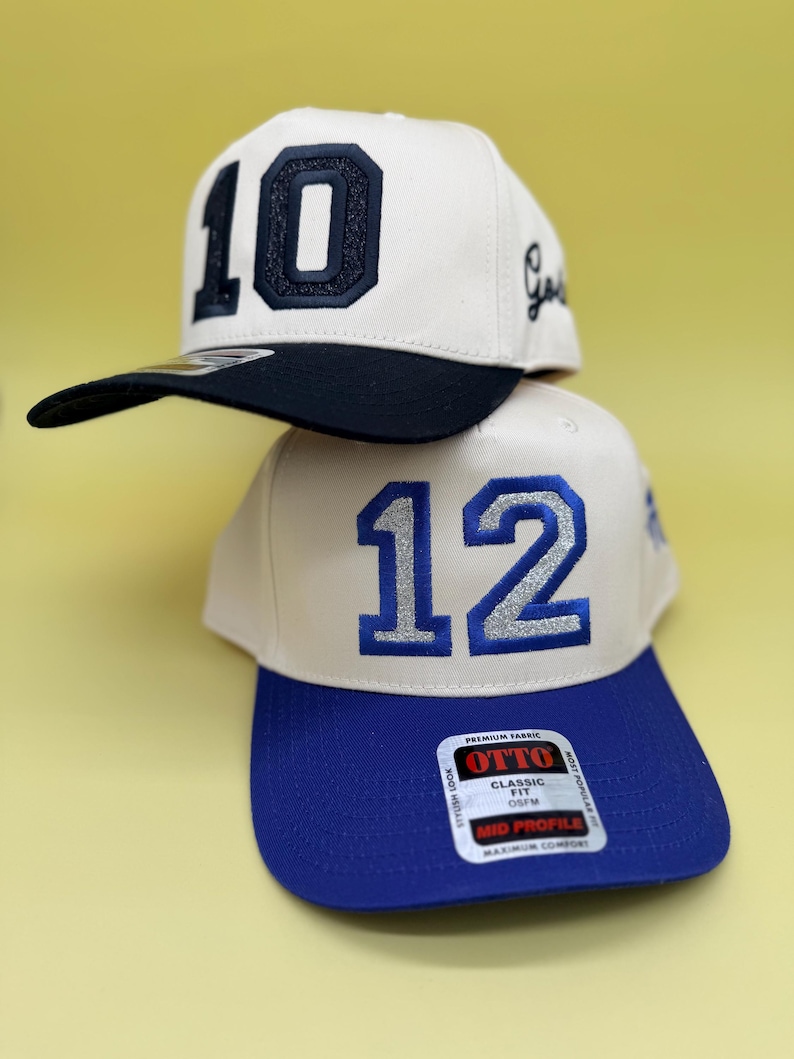 Puede incluir: Dos gorras de b&eacute;isbol apiladas verticalmente. La gorra superior es de color blanco roto con visera negra y el n&uacute;mero 10 en purpurina negra. La gorra inferior es de color blanco roto con visera azul y el n&uacute;mero 12 en purpurina azul, con una pegatina de la marca OTTO.
