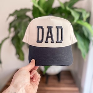 Puede incluir: Una gorra de b&eacute;isbol blanca con visera negra y la palabra "DAD" bordada en negro en la parte delantera.