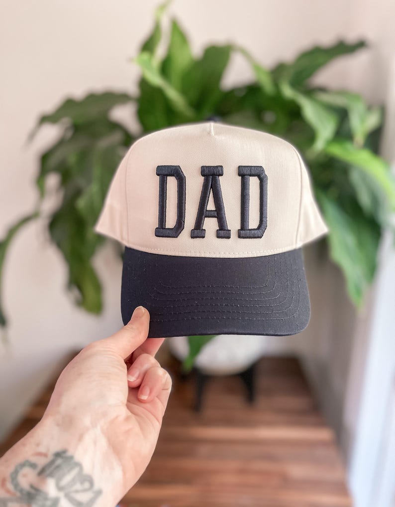 Puede incluir: Una gorra de b&eacute;isbol blanca con visera negra y la palabra "DAD" bordada en negro en la parte delantera.