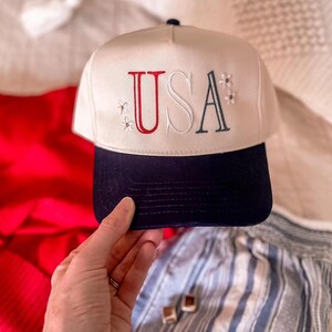 May include: A cream-colored baseball cap with a navy blue brim. The word "USA" is embroidered on the front in red and navy blue, with small floral accents. The cap is a patriotic accessory.