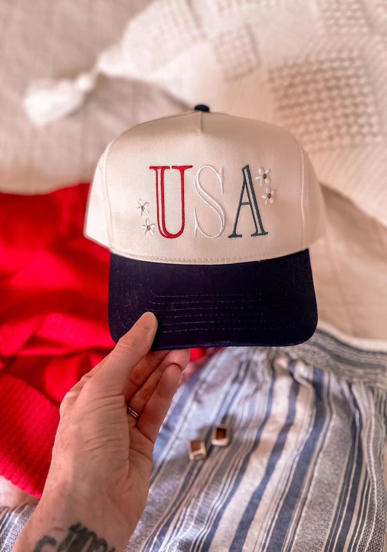 May include: A cream-colored baseball cap with a navy blue brim. The word "USA" is embroidered on the front in red and navy blue, with small floral accents. The cap is a patriotic accessory.