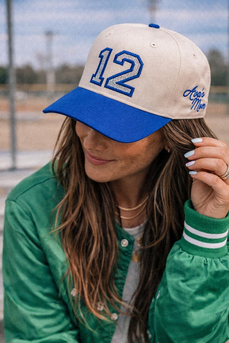 Puede incluir: Una gorra de b&eacute;isbol beige con visera azul brillante y un bot&oacute;n azul en la parte superior. El frente presenta el n&uacute;mero 12 en purpurina plateada con un borde azul. El lateral tiene el texto Ava's Mom bordado en letras azules.