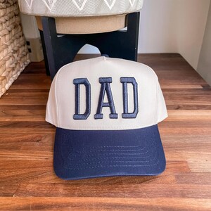 Puede incluir: Una gorra de b&eacute;isbol blanca con una visera azul marino y la palabra "DAD" bordada en azul marino en la parte delantera.