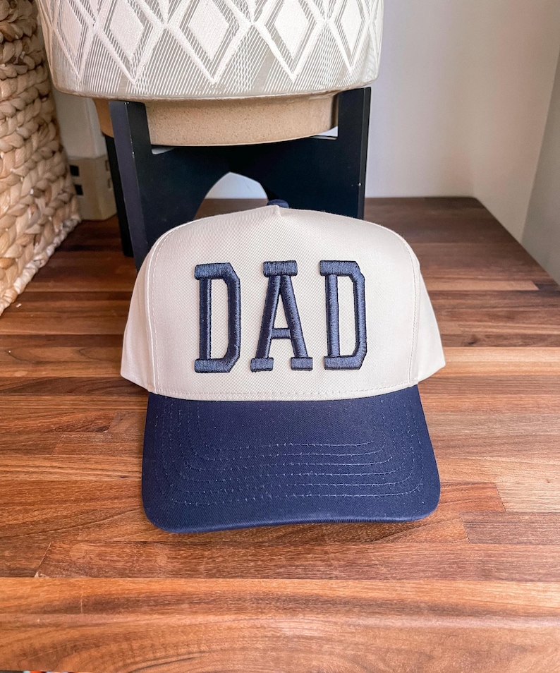 Puede incluir: Una gorra de b&eacute;isbol blanca con una visera azul marino y la palabra "DAD" bordada en azul marino en la parte delantera.