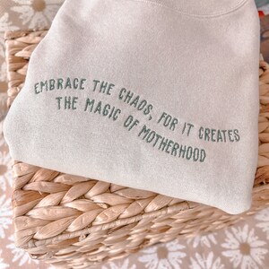 May include: A beige sweatshirt with the text "Embrace the chaos, for it creates the magic of motherhood" embroidered in green.