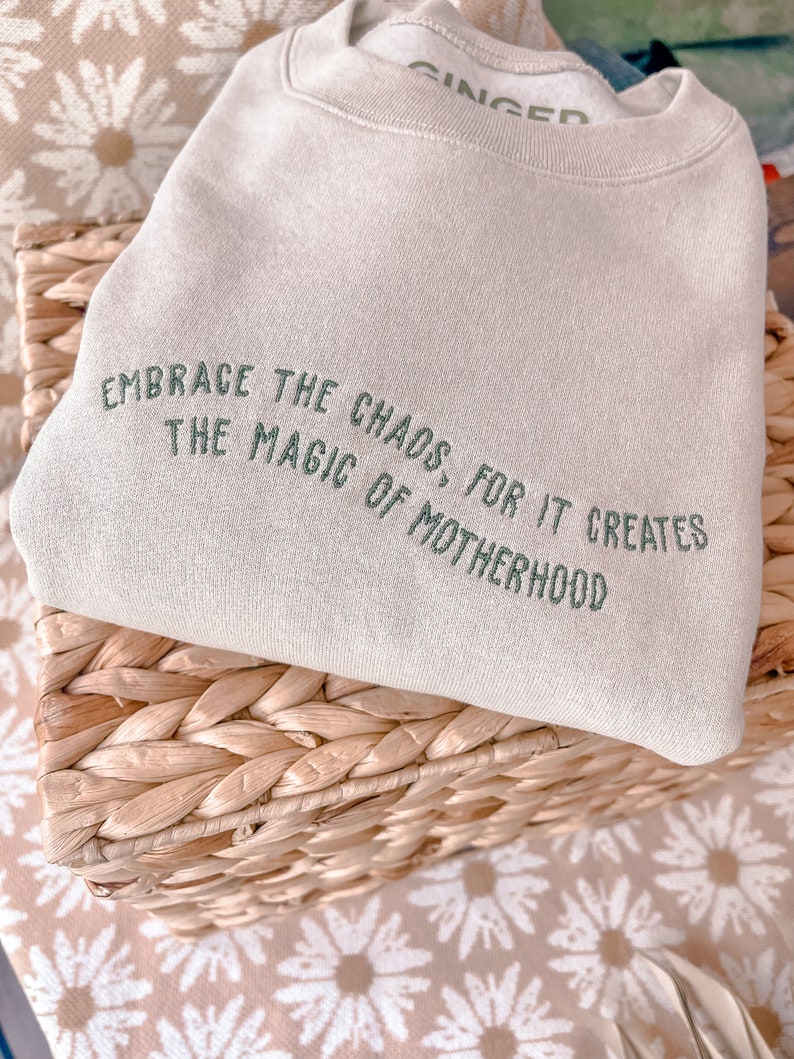 May include: A beige sweatshirt with the text "Embrace the chaos, for it creates the magic of motherhood" embroidered in green.