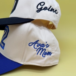 Puede incluir: Dos gorras de b&eacute;isbol de color blanco roto apiladas verticalmente. La gorra superior tiene una visera negra y el texto bordado "Goins" en letra cursiva negra. La gorra inferior tiene una visera azul y el texto "Ava's Mom" bordado en azul.