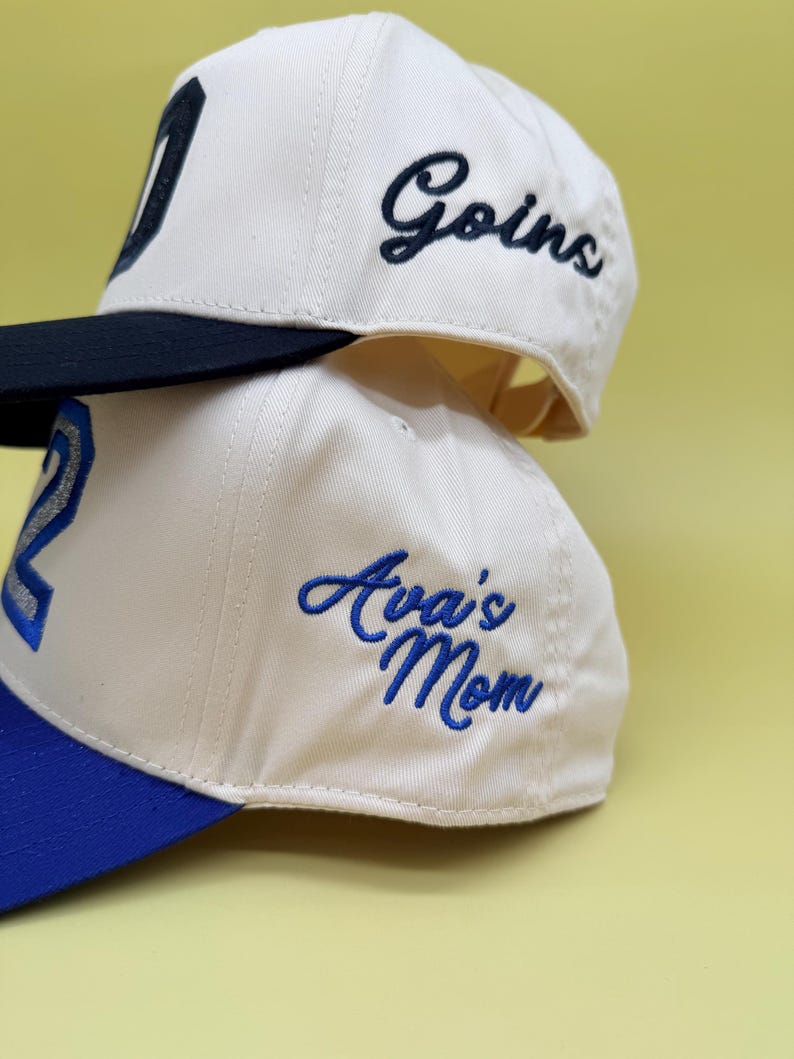 Puede incluir: Dos gorras de b&eacute;isbol de color blanco roto apiladas verticalmente. La gorra superior tiene una visera negra y el texto bordado "Goins" en letra cursiva negra. La gorra inferior tiene una visera azul y el texto "Ava's Mom" bordado en azul.