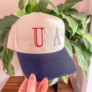 May include: A beige baseball cap with a navy blue brim. The word "USA" is embroidered on the front in red and teal, with small floral accents. The cap is held in front of a green leafy plant.