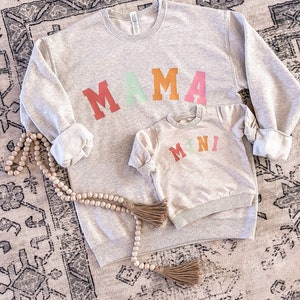 K&ouml;nnte beinhalten: Ein graues Sweatshirt mit dem Wort "MAMA" in bunten Blockbuchstaben. Ein kleineres graues Sweatshirt mit dem Wort "MINI" in bunten Blockbuchstaben liegt auf dem gr&ouml;&szlig;eren Sweatshirt.