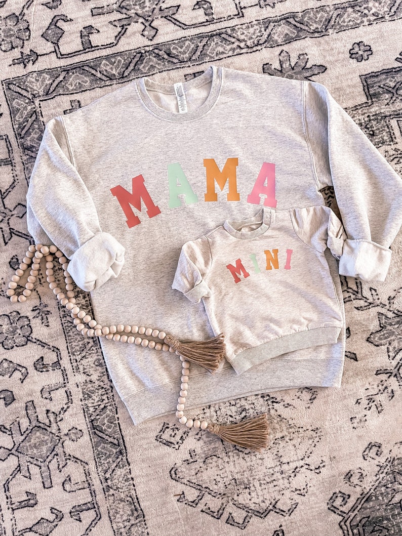 K&ouml;nnte beinhalten: Ein graues Sweatshirt mit dem Wort "MAMA" in bunten Blockbuchstaben. Ein kleineres graues Sweatshirt mit dem Wort "MINI" in bunten Blockbuchstaben liegt auf dem gr&ouml;&szlig;eren Sweatshirt.