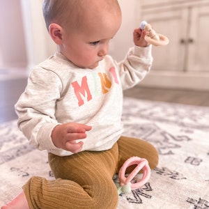 K&ouml;nnte beinhalten: Ein Baby tr&auml;gt ein hellgraues Sweatshirt mit dem Wort "MIRT" in Regenbogenfarben bedruckt und braune gerippte Leggings. Es sitzt auf einem wei&szlig;en und grauen Teppich. Das Baby h&auml;lt einen Holz-Bei&szlig;ring mit einer blauen Perle und einem rosa Ring.