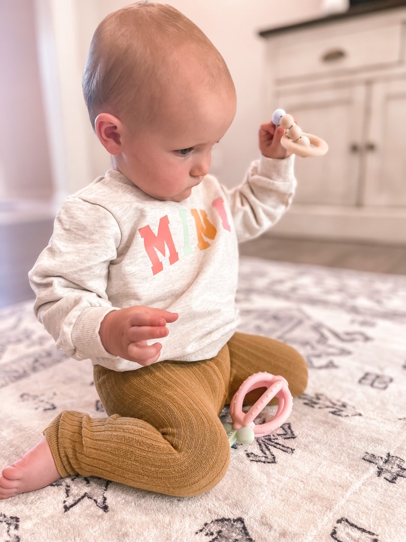 K&ouml;nnte beinhalten: Ein Baby tr&auml;gt ein hellgraues Sweatshirt mit dem Wort "MIRT" in Regenbogenfarben bedruckt und braune gerippte Leggings. Es sitzt auf einem wei&szlig;en und grauen Teppich. Das Baby h&auml;lt einen Holz-Bei&szlig;ring mit einer blauen Perle und einem rosa Ring.