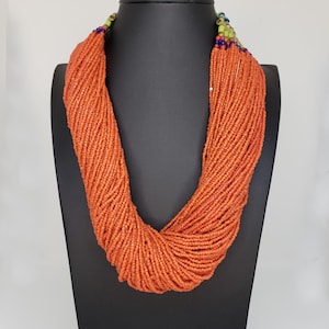 Puede incluir: Un collar largo de múltiples hilos hecho de pequeñas cuentas de color naranja. El collar se muestra en un busto de maniquí negro.