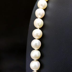 May include: A strand of white pearls on a black display stand. The pearls are round and smooth, and they are strung together on a thin, white thread.