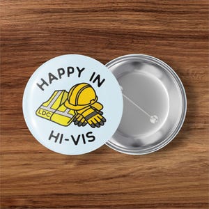 May include: A light blue button with the words "HAPPY IN HI-VIS" and a graphic of a yellow hard hat, gloves, and a safety vest. The button is next to a silver button.