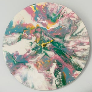 Puede incluir: Un reloj redondo con fondo blanco y patrones de pintura rosa, verde y azul en espiral. Las manecillas del reloj son doradas.