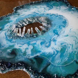 Op de afbeelding: Een blauw en wit kunsthars kunstwerk met een helder kristal centrum. Het hars is gezwirld en heeft een geode-achtige uitstraling. Het stuk is omgeven door kleine blauwe en heldere glazen stukjes.