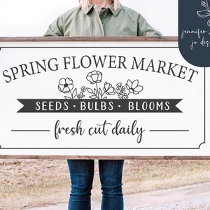 May include: A white wooden sign with black text that reads "Spring Flower Market" with a banner underneath that says "Seeds - Bulbs - Blooms" and the text "fresh cut daily" below the banner. The sign is being held by a person in front of a field.