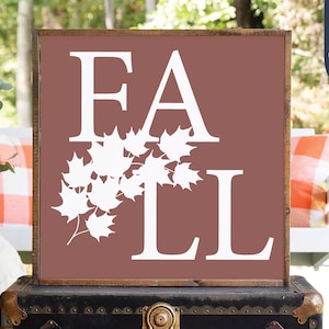 May include: A wooden sign with a brown background and white lettering that reads "FALL". The letters are decorated with white maple leaves.