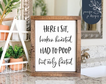 May include: A white wooden sign with black text that reads "HERE I SIT, broken hearted. HAD TO POOP but only farted." The sign is sitting on a countertop in a bathroom.