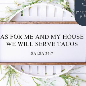 Pode incluir: Uma placa de madeira branca com moldura marrom, com o texto "As for me and my house we will serve tacos" e "Salsa 24:7". A placa é cercada por flores brancas e vegetação.