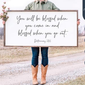 May include: A framed wooden sign with the text "You will be blessed when you come in and blessed when you go out. Deuteronomy 28:6" in a handwritten font. The sign is held by a person wearing a green jacket and blue jeans.