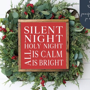 May include: A red wooden sign with white text that reads "Silent Night Holy Night All Is Calm Is Bright". The sign is surrounded by greenery and red berries.