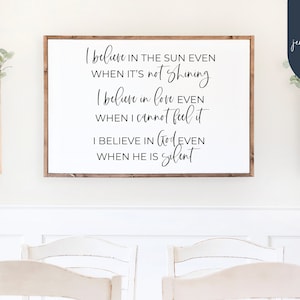 May include: A white wooden sign with black calligraphy text that reads "I believe in the sun even when it's not shining. I believe in love even when I cannot feel it. I believe in God even when he is silent."