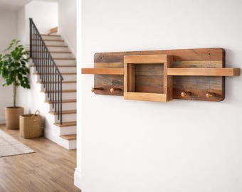 May include: A rustic wooden wall shelf with a rectangular backboard, two shelves, a central square cubby, and three wooden pegs. The shelf is made of various wood tones and is mounted on a white wall. The design is suitable for entryway or hallway decor.