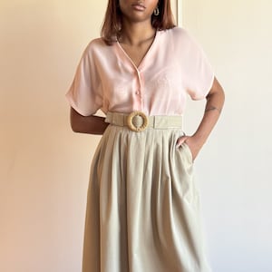 May include: A light pink, sheer blouse with a v-neck and short sleeves. The blouse is paired with a high-waisted, pleated, beige skirt and a brown woven belt with a large, round buckle.