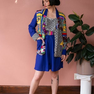 May include: A woman wearing a blue dress with a colorful patterned jacket and black cowboy hat. She is wearing black ankle boots and has a tattoo on her leg.