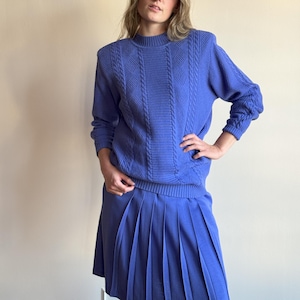 Puede incluir: Un jersey azul de punto de cable y una falda plisada azul a juego. El jersey tiene cuello alto y mangas largas. La falda es de largo hasta la rodilla y tiene una cintura ajustada.