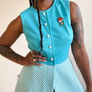 May include: A sleeveless blue dress with a white polka dot pattern. The dress has a button-up front and a mushroom-shaped brooch.