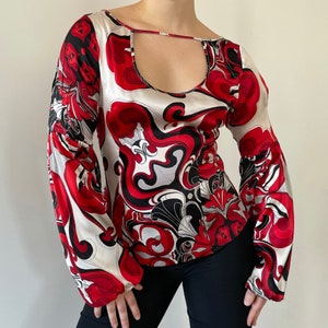 Puede incluir: Una blusa de manga larga blanca, roja y negra con un patrón geométrico. La blusa tiene un escote redondo y un recorte de ojo de cerradura en el escote.