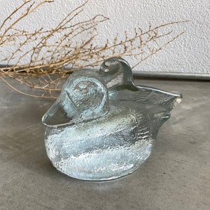 Puede incluir: Una figurita de pato de vidrio transparente con una superficie texturizada. El pato está sentado sobre una superficie gris.