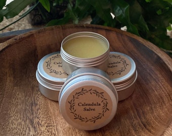 May include: Three small metal tins of Calendula Salve, stacked on a wooden surface. The top tin is open, revealing a yellow salve.
