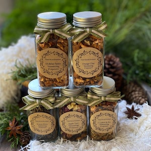 May include: Five glass jars filled with dried fruit and herbs, each with a brown paper label with a wreath design and text that reads "The Room Boutique" and the name of a simmering potpourri scent: "Holiday Spice", "Orange Chai", "Forest Floor", "Apple Tart", and "Comfort & Joy".