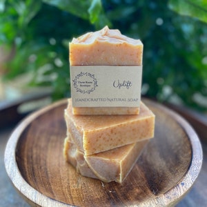 Puede incluir: Tres barras de jabón natural artesanal apiladas en una bandeja de madera. La barra superior tiene una etiqueta de papel marrón que dice "Uplift" y "Three Roots Boutique".