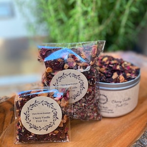 May include: Two clear plastic bags filled with loose leaf fruit tea. The tea is a mix of dried fruit pieces, flowers, and herbs. The bags are labeled "Three Roos Boutique Cherry Vanilla Fruit Tea" and "Three Roos Boutique Vanilla Tea".