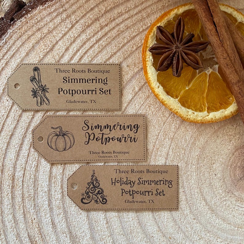 Puede incluir: Una composici&oacute;n r&uacute;stica con tres etiquetas de papel marr&oacute;n para juegos de popurr&iacute; para hervir a fuego lento. Palitos de canela, an&iacute;s estrellado y una rodaja de naranja seca acompa&ntilde;an las etiquetas, todo sobre una superficie de madera.