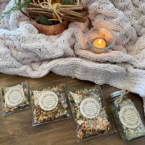 May include: Four clear plastic bags filled with a blend of dried fruits, herbs, and spices. The bags are labeled "Autumn Woods Simmering  " with a decorative wreath design. A glass jar filled with the same blend is also pictured.
