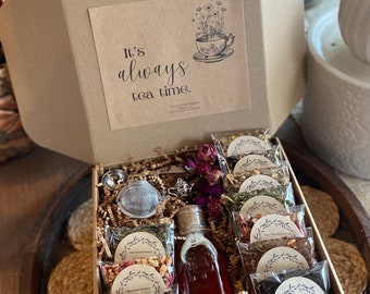May include: A brown cardboard box filled with tea bags and a bottle of honey. The tea bags are labeled with different flavors, including "Palais Royal Blackberry Fruit Tea", "Earl Grey Black Tea", and "Three Blooms Sampler Green Apple Cinnamon". The box also includes a tea strainer and a small card that says "It's always tea time."