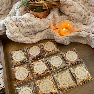 May include: Twelve small clear plastic bags filled with dried herbs and fruit peels. Each bag has a brown paper circle label with the text "Timeless Blends" and the name of a tea blend. The blends include "Autumn Spice", "Apple Tart", "Orange Chai", "Sweet Lemon", "Cranberry Ginger", "Lemon Lavender", "Herbal Citrus", "Autumn Spice", "Orange Hibiscus", "Forest Floor", "Lemon & Ginger", and "Earl Grey & Orange".