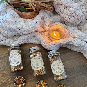 May include: Three glass jars filled with popcorn and labeled with different scents: "Comfort & Joy", "Orange Oat", and "Signature Spice". The jars are on a wooden surface with a white knitted blanket and a lit candle in the background.