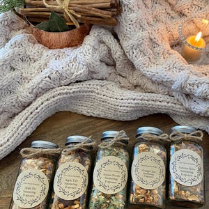 May include: Five glass jars with brown paper labels and twine tied around the lids. Each jar contains a different type of simmering potpourri. The labels read: "Apple Pie Simmering Potpourri", "Apple Tart Simmering Potpourri", "Autumn Woods Simmering Potpourri", "Orange Chai Simmering Potpourri", and "Autumn Spice Simmering Potpourri".