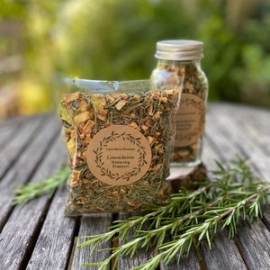 May include: A clear glass jar with a silver lid filled with dried herbs and fruit. The jar is labeled "Lemon-licious Simmering Potpourri". The jar is sitting on a wooden table next to a clear plastic bag filled with the same potpourri.