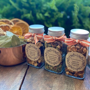 May include: Three glass jars filled with dried fruit and herbs, tied with orange and white ribbon. The jars are labeled "Apple Tart Simmering Potpourri", "Autumn Woods Simmering Potpourri", and "Apple Pie Simmering Potpourri".