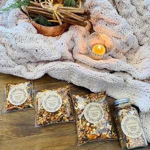 May include: A cozy scene featuring a cream-colored cable-knit blanket, a copper cup with cinnamon sticks and dried orange slices, and several packages and a jar of Orange Chai Seasoning. A lit candle adds warmth.